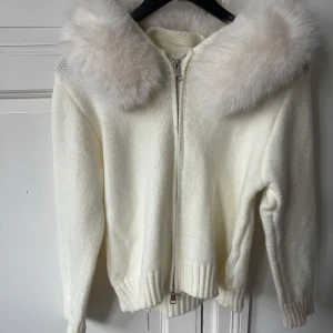 Vit stickad kofta med pälskrage - Säljer en elegant vit stickad kofta med en lyxig pälskrage. Koftan har en dragkedja framtill och långa ärmar. Perfekt för kyliga dagar när du vill känna dig både varm och stilren.