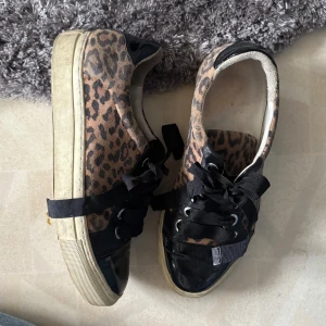Leopardmönstrade sneakers med svarta detaljer - Snygga sneakers med leopardmönster och svarta detaljer. Skorna har svarta snören och en vit sula. Perfekta för att ge en trendig touch till din outfit.