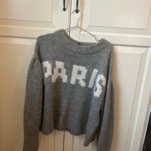 Grå stickad tröja från H&M - Säljer en grå stickad tröja från H&M med texten 'PARIS' i vitt på framsidan. Tröjan har en rund halsringning och långa ärmar. Har själv storlek M men ville ha en större passform då den var croped