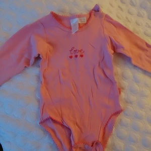 Rosa långärmad body med hjärtan, 6-9 månader  - Söt rosa body från Pumpkin Patch med texten 'love' och tre små hjärtan broderade på framsidan. Den har långa ärmar och knäppning i grenen för enkel påklädning. Perfekt för små barn som vill ha något bekvämt och gulligt.