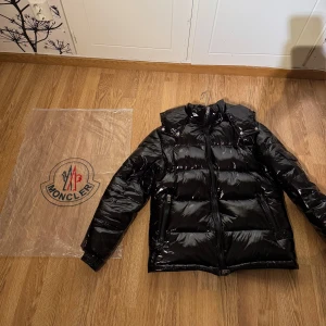 Moncler Maya jacka  - Säljer en svart Moncler Maya jacka med glansig finish. Jackan har en dragkedja framtill och långa ärmar. Är i väldigt bra skick enbart testad men aldrig använd, säljer då den är för liten för mig. Den är i storlek 0/XS men passar också smal, skriv privat om du har några frågor eller funderingar.