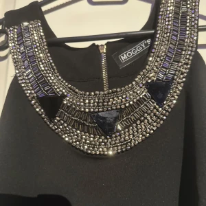 Svart klänning med pärldetaljer från Moggy's - Elegant svart klänning med imponerande pärldetaljer runt halsringningen. Klänningen har en dragkedja i ryggen och är ärmlös, vilket ger en stilren och sofistikerad look. Perfekt för speciella tillfällen.