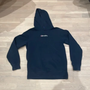 Mörkblå hoodie från Champion - Säljer en svart hoodie från Champion med broderad logga på bröstet. Den har en klassisk design med huva och långa ärmar. Perfekt för en avslappnad stil.  Storleken står i barn storlek så det liknar S/XS i storleken