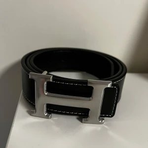 Svart bälte med silverfärgat spänne - Snyggt svart bälte i skinn med ett stort, silverfärgat H-format spänne. Perfekt för att ge en stilren touch till din outfit. Bältet har en elegant design med vita sömmar längs kanterna.