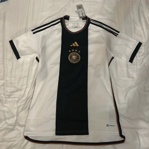 Tyskland hemma-tröja 2022 - Tysklands hemma-ställ från VM 2022. Tröjan är helt ny och har lapparna kvar. Tröjan är köpt på classicfpotballshirts.com, och är alltså helt äkta.
