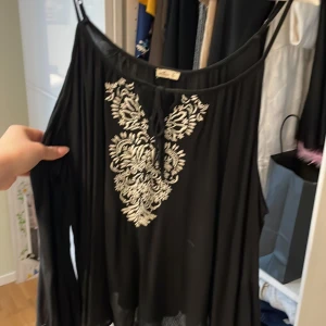 Svart blus från Hollister - Snygg svart blus från Hollister med vita broderade detaljer framtill. Blusen har långa ärmar och en luftig passform med knytning vid halsen. Perfekt för en stilren look.