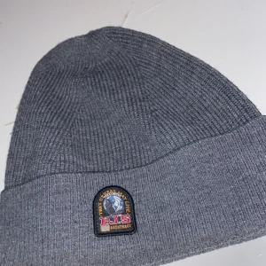 Parajumper mössa - Snygg grå ribbad mössa från Parajumpers med ett broderat märke framtill. Perfekt för kyliga dagar och ger en stilren look. Mössan är mjuk och bekväm att bära.