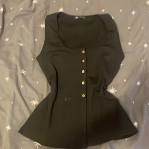 Svart top från Zara - Snygg svart top från Zara med knappar framtill. Den är ärmlös och har en elegant passform som framhäver figuren. Perfekt för en stilren look. 