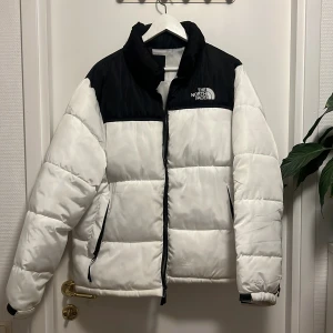 Vit och svart pufferjacka från The North Face - Säljer en snygg vit och svart pufferjacka från The North Face. Jackan har en broderad logga på bröstet och ryggen, samt praktiska dragkedjefickor. Perfekt för kyliga dagar med sin varma och bekväma design. Storlek L men passar även XL, hör av er vid minsta fundering. Det är ett litet hål i vänster ficka men det är det ända.