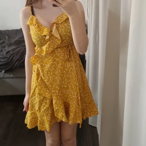 Gul blommig klänning - Söt gul klänning med vita blommor och volangdetaljer. Den har en omlott-design med knytning i midjan och smala axelband. Perfekt för sommarens varma dagar!
