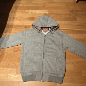 Grå hoodie från Burberry - Säljer en stilren grå hoodie från Burberry med dragkedja och klassiskt rutigt foder. Tröjan har en broderad logga på bröstet och är perfekt för en avslappnad look. Huvan är fodrad med det ikoniska Burberry-mönstret.