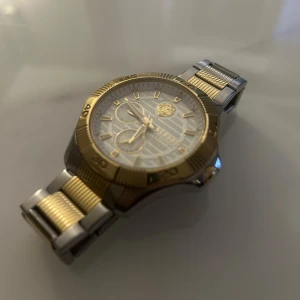 Versace originalklocka-Guld & silver, toppskick (pris kan diskuteras)  - Säljer en äkta Versace-klocka i guld och silver. Klockan har en vit urtavla med guldfärgade detaljer och två mindre visare. Armbandet är i rostfritt stål med guldpläterade länkar. Den är i väldigt fint skick, inga repor eller skador. En snygg och elegant klocka för den som gillar lyxig design.