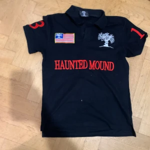 Haunted Mound x Sematary Polo - Snygg svart pikétröja från Haunted Mound med broderade detaljer. Tröjan har en stor röd broderad trädgrafik på baksidan och texten 'HAUNTED MOUND' på framsidan. Den har korta ärmar och en klassisk krage med knappar.