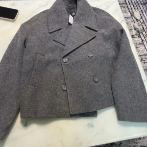 Grå kappa från 157 - Snygg grå kappa från 157 med dubbelknäppning och bred krage. Jackan har en stilren design och är perfekt för kyligare dagar. Den är tillverkad i ett mjukt material som ger en bekväm passform.