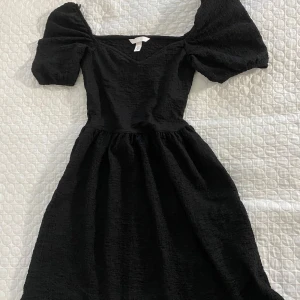 Svart klänning från H&M - Säljer en svart klänning från H&M med puffiga kortärmar och en v-ringad hals. Klänningen har en texturerad yta och en smickrande passform som går ner till knäna. Perfekt för en stilren look!