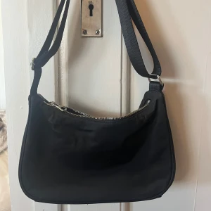 Svart axelväska från Weekday i nylon med silverdetaljer - Snygg svart axelväska från Weekday med dragkedja upptill. Perfekt för att bära med sig det nödvändigaste. Stilren design som passar till många olika outfits. Genom att förlänga bandet kan väskan bäras som en crossbody väska