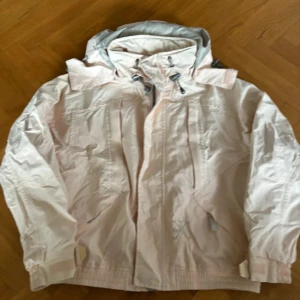 Kyra K jacka  - Säljer en ljusrosa kyra K vindjacka med justerbar huva och dragkedja framtill. Jackan har långa ärmar och praktiska fickor. Perfekt för blåsiga dagar och håller dig skyddad mot väder och vind. Helt ny 