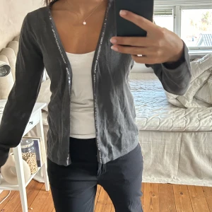 Grå kofta från Rabens Saloner - Snygg grå kofta från Rabens Saloner i storlek S. Koftan har långa ärmar och en glittrig kant längs med knäppningen. Perfekt för att lägga till lite bling i vardagen.