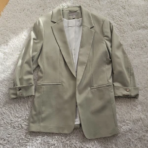 Pistage-grön kavaj från H&M - Snygg kavaj i ljusgrön / Pistage från H&M med trekvartsärmar och stilrena knappar i silver på ärmsluten. Perfekt för en chic look. Kavajen har en klassisk krage och två fickor framtill. Fickorna är bara för synes skull (igensydda) 