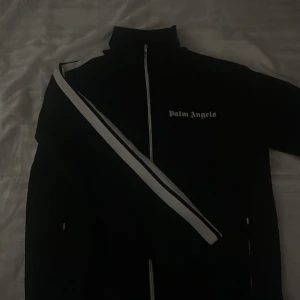 Svart track jacket från Palm Angels - Hej, säljer en svart track jacket från Palm Angels med vit dragkedja och vita ränder längs ärmarna. Jackan har en hög krage och Palm Angels-loggan tryckt på bröstet. Perfekt för en stilren och sportig look. Pris kan diskuteras!
