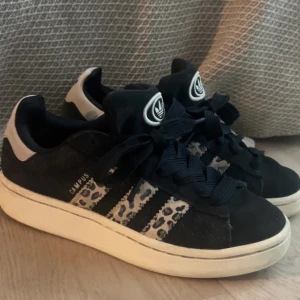 Svarta Adidas Campus sneakers med leopardmönster - Snygga svarta Adidas Campus sneakers med leopardmönstrade detaljer på sidorna. Skorna har en klassisk design med vita sulor och Adidas logga på plösen och hälen. Perfekta för en trendig look.