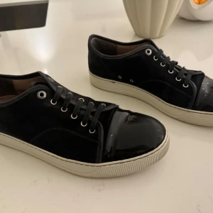 Mörkgrå sneakers från Lanvin - Snygga svarta sneakers från Lanvin med en kombination av mocka och lackdetaljer. Skorna har en vit sula och klassisk snörning. Perfekta för en stilren look. BOX (ingår)!