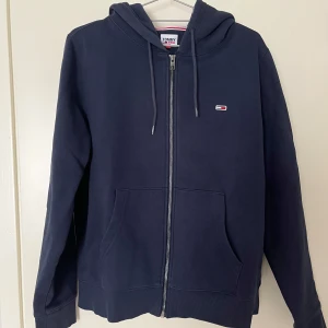 Mörkblå hoodie från Tommy Hilfiger Jeans - Säljer en stilren mörkblå hoodie från Tommy Hilfiger Jeans med dragkedja och justerbar huva. Tröjan har långa ärmar och en liten logotyp på bröstet. Perfekt för en avslappnad look.