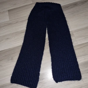 Mörkblå stickad halsduk - Säljer en stilren mörkblå stickad halsduk. Den är perfekt för att hålla dig varm under kyliga dagar. Halsduken har en enkel design med en mjuk och bekväm känsla. Måtten är cirka 120 cm i längd och 20 cm i bredd.