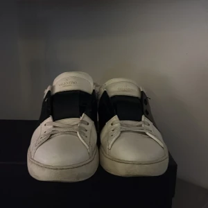 Valentino sneakers i vitt och svart - Snygga Valentino sneakers i vitt med en bred svart rem över snörningen. Skorna har en stilren design med diskreta nitar på hälen. Perfekta för en trendig look.priset kan diskuteras äkta!!