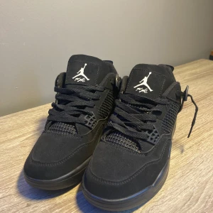Svarta Nike Air Jordan 4’s - Snygga svarta Nike Air Jordan 4’s med klassisk design och snörning. Skorna har en stilren look med Jumpman-loggan i vitt på tungan och hälen. Perfekta för dig som vill ha en stilren och sportig stil.