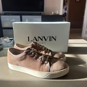 Bruna sneakers från Lanvin - Snygga bruna sneakers från Lanvin i mocka med snörning. Skorna har en stilren design med en vit sula och är perfekta för en avslappnad look. Skadorna på skorna visas i bilderna, har du frågor eller vill ha mer bilder är det bara att skriva till mig! Dustbag och nya skosnören medföljer🙌🏼