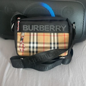 Rutig axelväska från Burberry - Snygg axelväska från Burberry med klassiskt rutigt mönster i beige, svart och rött. Väskan har en justerbar axelrem och en praktisk dragkedja. Perfekt för att bära dina nödvändigheter med stil. Kan även diskutera priset 