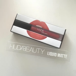 Huda Beauty Liquid Matte Läppstift i nyansen Bombshell - Helt ny, aldrig använd. Säljer ett flytande matt läppstift från Huda Beauty i nyansen Bombshell. Denna färg är en vacker nude-rosa ton som ger en elegant och långvarig finish. 