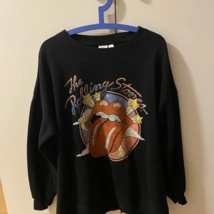 Svart Rolling Stones sweatshirt - Säljer en svart sweatshirt med Rolling Stones-tryck. Tröjan har ett stort färgglatt tryck av den ikoniska tungan och texten 'The Rolling Stones' i glittriga bokstäver. Perfekt för fans av bandet!