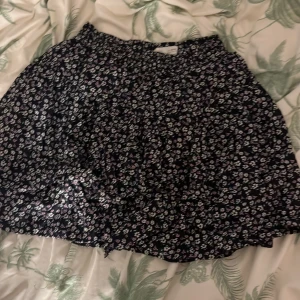 Blommig kortkjol - Söt blommig kortkjol i mörk färg med små vita och lila blommor. Perfekt för en avslappnad stil. Resår i midjan för bekväm passform.