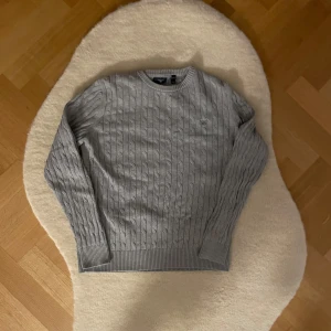 Grå kabelstickad tröja från GANT (164) - Säljer en stilren grå kabelstickad tröja från GANT. Tröjan har en rund halsringning och ett diskret märke på bröstet. Perfekt för kyligare dagar med sin klassiska design och bekväma passform. Storleken är 158-164 cm och motsvarar åldern 13/14. Vid frågor eller intresse vänligen dm mig!