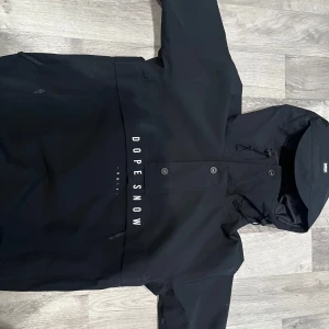 Svart jacka från Dope Snow - Säljer en svart jacka från Dope Snow med huva och tryckknappar. Jackan har en stilren design med logotypen 'DOPE SNOW' tryckt på framsidan och baksidan. Perfekt för kyliga dagar och ger en cool look. Skriv om ni vill ha fler bilder