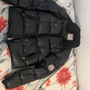 Svart dunjacka från Moncler - Säljer en snygg svart dunjacka från Moncler. Jackan har en quiltad design med dragkedja framtill och en logotyp på ärmen. Perfekt för kyliga dagar med sin varma och stilrena look.