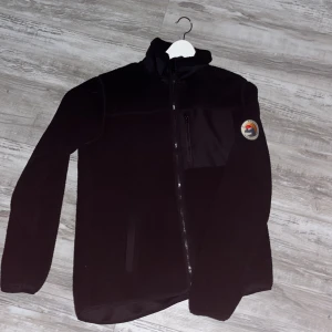 Svart fleecejacka med emblem - Säljer en svart fleecejacka med dragkedja och ett coolt emblem på ärmen. Jackan har en bröstficka med dragkedja och långa ärmar. Perfekt för kyliga dagar!