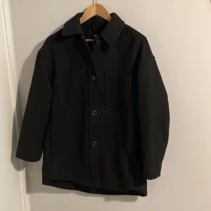 Svart jacka/kappa från H&M - Säljer en stilren svart jacka från H&M. Jackan har en klassisk design med knappar framtill och långa ärmar. Perfekt för höst och vår. Den är tillverkad i ett material som ger en snygg och modern look.