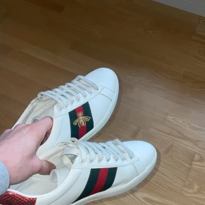 Vita sneakers med röd och grön detalj - Hej säljer nu mina snygga vita sneakers från Gucci med ikoniska röd-gröna ränder och en broderad bi-detalj på sidan. Skorna har snörning och en kontrasterande häl i rött och grönt. 