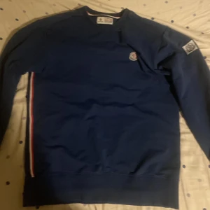 Blå tröja från Moncler - Säljer en snygg blå tröja från Moncler med deras ikoniska logga på bröstet och ärmen. Tröjan har en ribbad krage och ränder i rött, vitt och blått längs sidan. Perfekt för en stilren look.