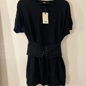 Svart klänning med bälte från Bik Bok - Snygg svart klänning från Bik Bok med korta ärmar och ett brett bälte med snörning i midjan. Perfekt för en stilren look. Klänningen har en enkel design som gör den lätt att matcha med olika accessoarer. Har aldrig använt så är helt ny!💗