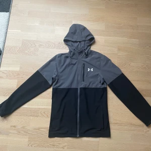 Under Armour jacka herr - Säljer en grå och svart jacka från Under Armour med huva och dragkedja. Jackan har en stilren design med en liten logotyp på bröstet. Perfekt för träning eller vardagsbruk. Storlek S 