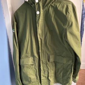 Grön jacka från Superdry st S  - Säljer en grön jacka från Superdry med huva och stora fickor framtill. Jackan har en cool patch på ärmen och stängs med knappar och dragkedja Perfekt för höst och vår!