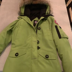 Grön parkajacka från Canada Goose - Säljer en grön parkajacka från Canada Goose med pälsdetaljer på huvan. Jackan har dragkedja och knappar framtill samt flera praktiska fickor. Perfekt för kalla dagar. 