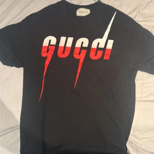 Svart t-shirt från Gucci - Snygg svart t-shirt från Gucci med ett iögonfallande rött och vitt logotyptryck på framsidan. Perfekt för en sommar dag , sticker ut men skit snygg . Inga skador .Har tyvärr inget kvitto därav billigt pris och använder aldrig den 