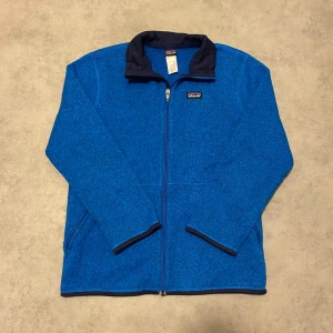 Blå fleecejacka från Patagonia - Säljer en snygg blå fleecejacka från Patagonia. Jackan har en dragkedja framtill och en hög krage i mörkare blå nyans. Perfekt för kyliga dagar och har en stilren design med två fickor framtill.