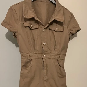 Beige kortärmad jeans/ denim  klänning med knappar - Snygg beige klänning med korta ärmar och hjärtformade knappar. Den har en klassisk krage och praktiska fickor framtill. Perfekt för en avslappnad stil.