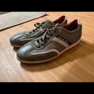 Grå sneakers från Hugo Boss - Snygga grå sneakers från Hugo Boss med vita och svarta detaljer. Skorna har snörning och en diskret logotyp på sidan. Perfekta för en stilren look.